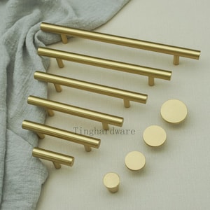 May include: A collection of gold-toned cabinet hardware, including bar pulls and round knobs. The bar pulls vary in length, and the knobs are circular. The hardware is arranged on a neutral-colored fabric. The text "Tinghardware" is visible.