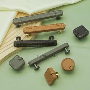 May include: Cabinet hardware featuring leather-wrapped handles and knobs in grey, brown, and black. The handles have silver or gold posts and white stitching. Knobs are square or round with matching stitching.