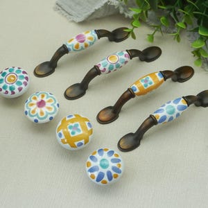 May include: A collection of decorative cabinet hardware, including handles and knobs. The handles have a dark bronze finish and are adorned with colorful floral ceramic designs. The knobs are round with matching floral patterns in various colors.