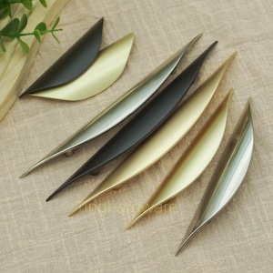 Op de afbeelding: Een verzameling moderne kastgrepen in diverse metallic afwerkingen, waaronder zwart, goud en zilver. De grepen hebben een strak, gebogen ontwerp, dat lijkt op een blad. De hardware is gerangschikt op een neutraal gekleurd stoffen oppervlak.