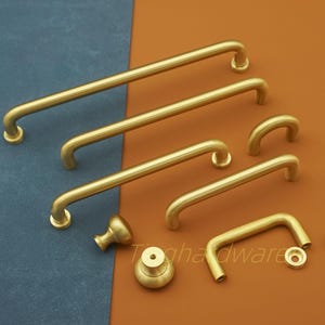 Puede incluir: Un conjunto de herrajes de latón para armarios, que incluye tiradores de barra de varios tamaños, un pomo y un tirador pequeño. Los herrajes están dispuestos sobre una superficie azul y marrón, mostrando los diferentes diseños y tamaños.