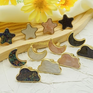 May include: A collection of decorative cabinet knobs in star, moon, and cloud shapes. The knobs feature gold-toned bases with colorful, textured inlays in shades of blue, white, pink, and black. The knobs are arranged on a wooden surface.