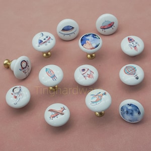 May include: A collection of white ceramic cabinet knobs with brass bases. Each knob features a different space-themed design, including rockets, planets, astronauts, and flying saucers. The knobs are round and have a glossy finish, perfect for adding a whimsical touch to furniture.