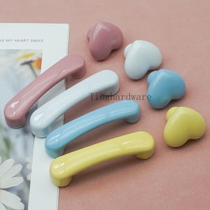 Op de afbeelding: Een verzameling kleurrijke keramische kastbeslagen, waaronder hartvormige knoppen en gebogen handgrepen. De handgrepen zijn in pastelroze, wit, lichtblauw en geel. De hartknoppen zijn roze, wit, lichtblauw en geel. De tekst "Tinghardware" is zichtbaar.