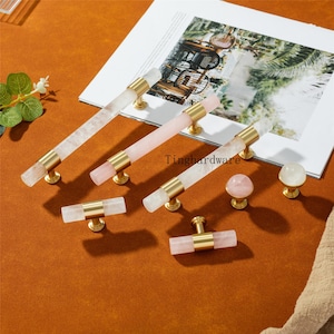 Op de afbeelding: Een verzameling decoratieve meubelbeslag, waaronder kristallen en rozenkwarts handgrepen en knoppen. De handgrepen hebben goudkleurige bases en accenten. De knoppen zijn bolvormig en paddenstoelvormig. Op een bruin oppervlak.