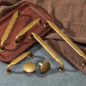 Pode incluir: Uma coleção de ferragens de armário de latão antigo, incluindo puxadores e botões. Os puxadores têm um design curvo e são montados em bases redondas. O hardware é exibido em uma bandeja de madeira e um fundo de tecido marrom.