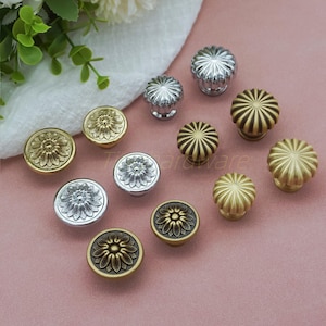 May include: A selection of decorative cabinet knobs in gold, silver, and bronze finishes. The knobs showcase floral and geometric patterns, with diverse textures and shapes. The knobs are displayed on a pink surface. Measurements are in centimetres.