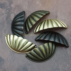 May include: A collection of decorative shell-shaped cabinet pulls in various metallic finishes. The pulls are in shades of bronze, brass, and black, with a ridged, fan-like design. The hardware is arranged on a neutral surface.