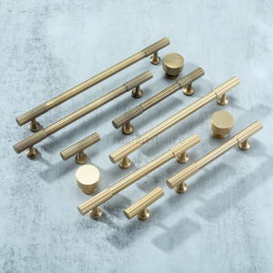 May include: A selection of brass-coloured cabinet handles and knobs. The bar handles have a grooved design, and the knobs are cylindrical. The hardware is displayed on a light grey surface. Measurements are in centimetres.
