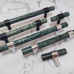 Op de afbeelding: Een verzameling groene marmeren kastgrepen met zwarte en zilveren hardware. De grepen zijn cilindrisch met een gemarmerd patroon en liggen op een wit marmeren oppervlak. De hardware omvat zwarte en zilveren metalen accenten.