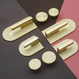 May include: A collection of gold-toned cabinet hardware, including handles and knobs. The handles have a rectangular bar design atop a curved, grooved base. The knobs are circular with a spiral pattern. The hardware is displayed on a brown and pink background.