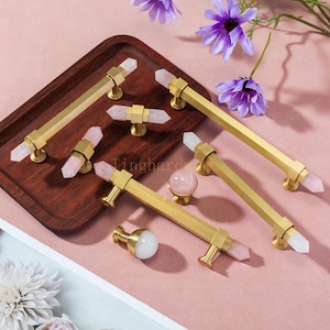 Peut inclure: Ensemble de poignées et de boutons de placard dorés avec des accents de cristal. Les poignées sont rectangulaires avec des cristaux à chaque extrémité. Les boutons sont ronds et cylindriques. Présentés sur un plateau en bois et une surface rose.