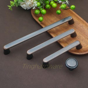 May include: A set of three cabinet pulls and a knob in varying sizes, with a dark grey finish and bronze accents. The hardware is displayed on a wooden tray, showcasing a modern design.