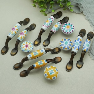 以下が含まれることがあります： 装飾的なキャビネットハードウェアのコレクション。ハンドルとノブが含まれています。ハンドルは、カラフルな花の模様が施された白いセラミックセンターを備えた、濃いブロンズ仕上げです。ノブは、お揃いのデザインの丸い形をしています。