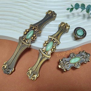 Op de afbeelding: Een assortiment meubelbeslag in antieke stijl in brons- en goudtinten. De set bevat twee lange handgrepen, een kleine handgreep en een knop, elk met een ovaal, lichtgroen stenen midden. Het beslag wordt gepresenteerd op een houten oppervlak.