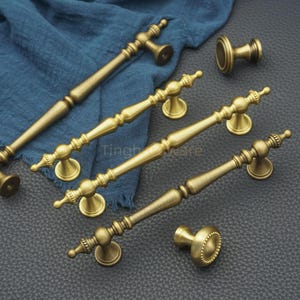May include: An assortment of antique brass cabinet pulls and knobs. The hardware features ornate detailing and a warm, golden finish. The pulls are various lengths, and the knobs have a round design. The hardware is displayed on a dark surface.
