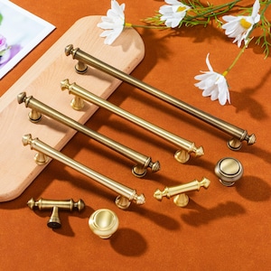 May include: An assortment of antique brass cabinet hardware, including long bar pulls, T-shaped pulls, and round knobs. The hardware is displayed on a wooden cutting board and a burnt orange surface, with white flowers in the background.