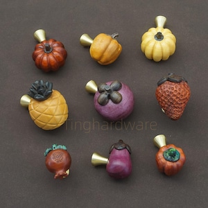 May include: A selection of decorative cabinet knobs shaped like fruits and vegetables. The knobs include a pumpkin, pineapple, strawberry, and aubergine, each with a brass stem. The knobs are in shades of orange, yellow, red, and purple.