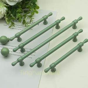 May include: A collection of sage green cabinet hardware, including several pull handles and two round knobs. The handles have a classic design with a central bar and decorative ends. The hardware is arranged on a white surface.