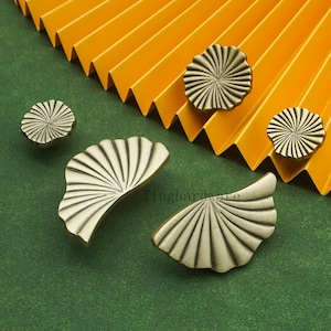 May include: A collection of antique brass cabinet hardware, including four round knobs and two fan-shaped pulls. The hardware features a radial design and is displayed on a green surface with a yellow paper fan in the background.