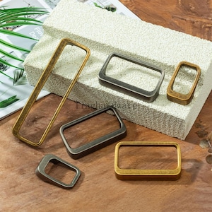 May include: An assortment of rectangular metal hardware pieces in various sizes and finishes, including gold, bronze, and gunmetal. The hardware is displayed on a textured, light-colored surface and a wooden surface. The image shows a variety of shapes and sizes.