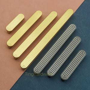 Op de afbeelding: Een verzameling gouden en zilveren kastgrepen met een groefontwerp. De grepen zijn rechthoekig met afgeronde uiteinden en zijn diagonaal gerangschikt. De gouden grepen zijn effen, terwijl de zilveren grepen witte strepen hebben. De tekst "Tinghardwa" is zichtbaar.