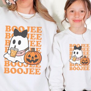 May include: Two white sweatshirts with a Halloween-themed graphic. The design features a ghost with a bow, holding a drink, and a jack-o'-lantern. The text "BOO JEE" is repeated in an orange font. One sweatshirt is worn by an adult, the other by a child.