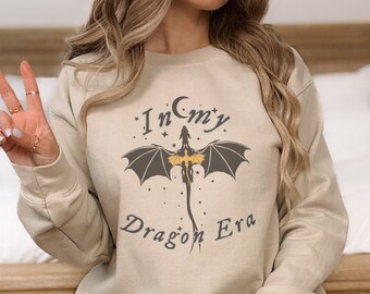 Puede incluir: Una sudadera beige con un gráfico de un dragón negro con detalles amarillos. El texto "In My Dragon Era" está impreso en la sudadera.