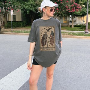 May include: Dark grey t-shirt with a tarot card design showing a skeleton holding an American flag and the words "THE PATRIOT." The shirt is worn with black shorts and a white cap. The image is taken outdoors.