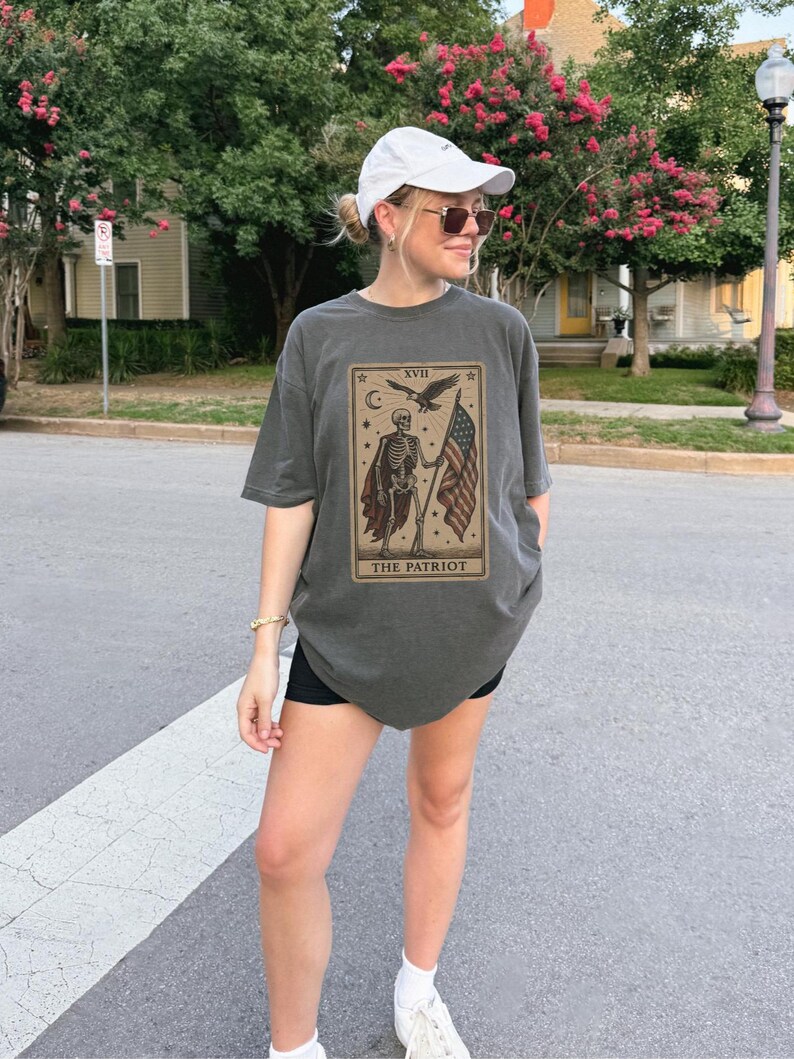May include: Dark grey t-shirt with a tarot card design showing a skeleton holding an American flag and the words "THE PATRIOT." The shirt is worn with black shorts and a white cap. The image is taken outdoors.