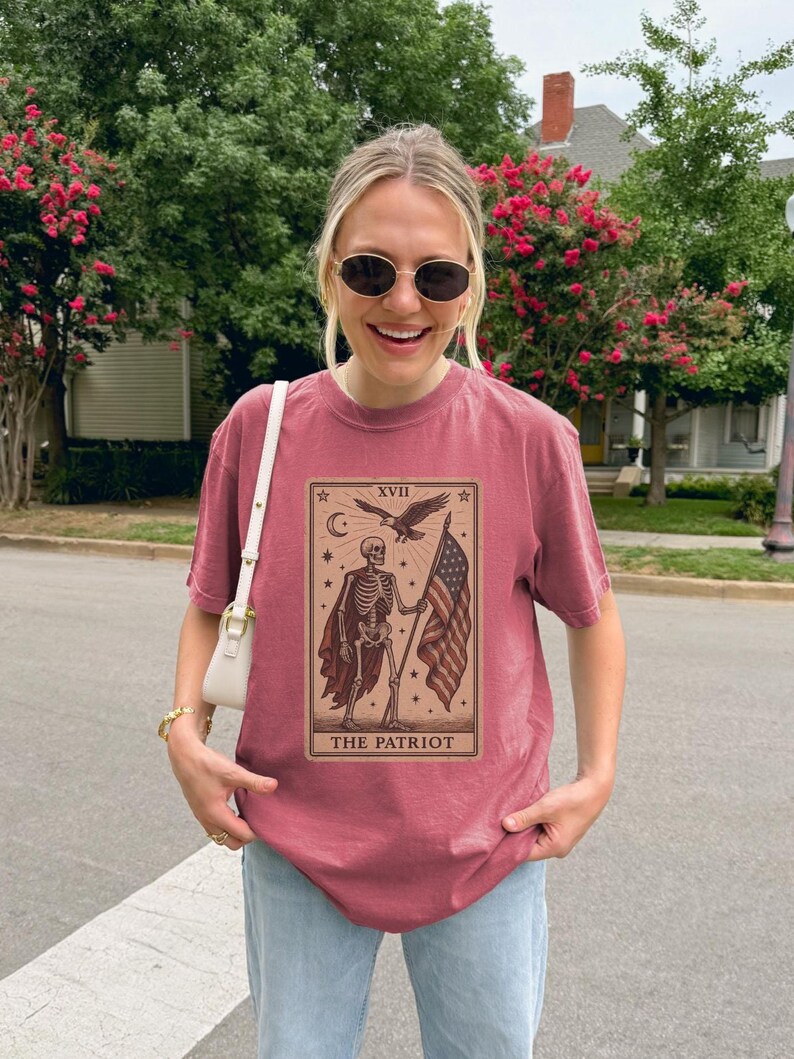 May include: A maroon t-shirt with a tarot card design. The design features a skeleton holding an American flag, an eagle, and the text "THE PATRIOT". The shirt is worn by a person wearing sunglasses.