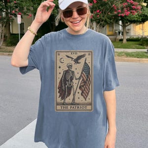 May include: A blue-grey t-shirt with a tarot card design. The card shows a skeleton holding an American flag, with an eagle and the text "THE PATRIOT". The shirt has a loose fit.