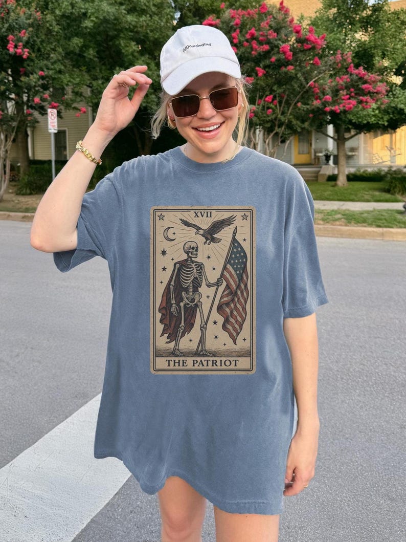 May include: A blue-grey t-shirt with a tarot card design. The card shows a skeleton holding an American flag, with an eagle and the text "THE PATRIOT". The shirt has a loose fit.