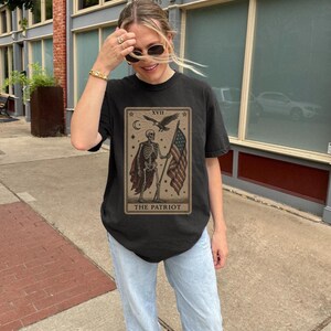 May include: Black t-shirt with a tarot card design depicting a skeleton holding an American flag. The card reads "XVII The Patriot" with stars, a crescent moon, and an eagle. The shirt is paired with light blue jeans.