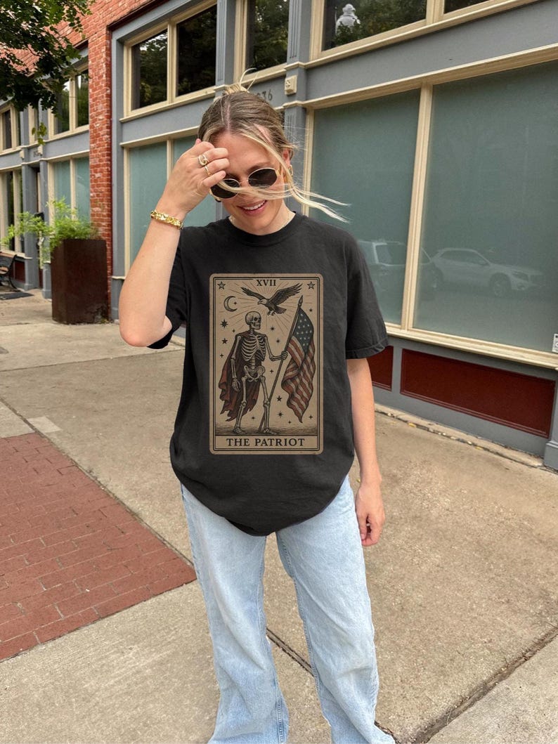May include: Black t-shirt with a tarot card design depicting a skeleton holding an American flag. The card reads "XVII The Patriot" with stars, a crescent moon, and an eagle. The shirt is paired with light blue jeans.