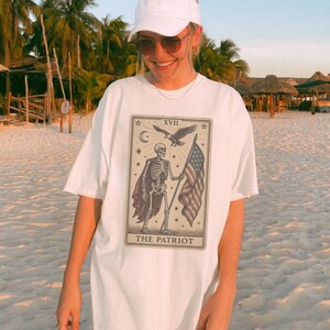 May include: White t-shirt with a tarot card design depicting a skeleton holding an American flag, an eagle, and the text "THE PATRIOT". The shirt is worn with a white cap and sunglasses.