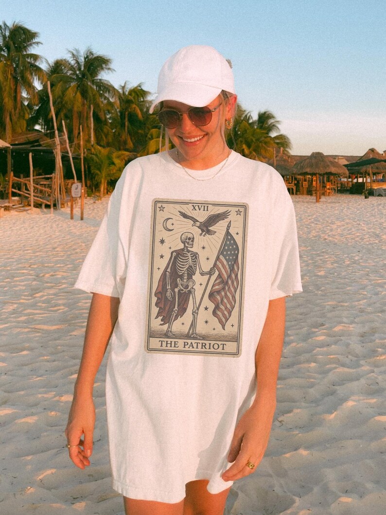May include: White t-shirt with a tarot card design depicting a skeleton holding an American flag, an eagle, and the text "THE PATRIOT". The shirt is worn with a white cap and sunglasses.