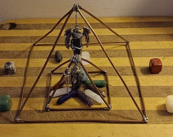 Op de afbeelding: Een koperen draadpiramide met een kristallen slinger die aan de bovenkant hangt. De slinger is een zwarte kristal met een zilveren drakenbedel. Er zijn zeven kristallen in de piramide, waaronder helder kwarts, amethist, rozenkwarts, groene aventurijn, lapis lazuli en rode jaspis.