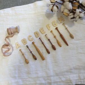 May include: A set of eight wooden bobbins for lace making, labelled with the letters A through H. The bobbins are arranged in a row on a white fabric background, with a spool of pink and white ribbon and a sprig of cotton bolls in the background.
