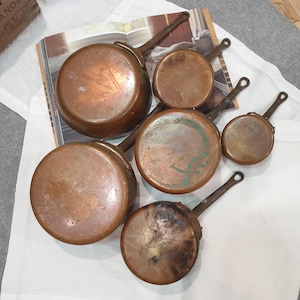May include: A set of six copper pans, each with a long, curved handle. The pans are of different sizes, with varying diameters. The copper surfaces display signs of age and use, with some areas darker. The handles are made of a darker metal.