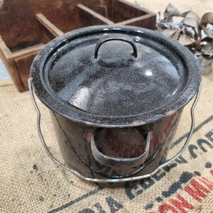 May include: A black enamel pot with a lid and a wire handle. The pot has a speckled pattern and a metal handle on the lid. The pot is sitting on a burlap surface.