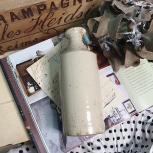 May include: An antique, off-white stoneware bottle with a narrow neck and a chipped rim. The cylindrical bottle has a glossy finish. It rests on a vintage book with handwritten notes and dried leaves.