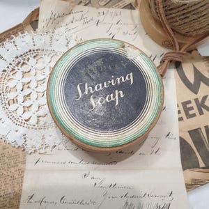 May include: A vintage shaving soap tin with a black and white label. The label reads "Shaving Soap". The tin is round with a green and white border. The background includes antique paper and lace, evoking a sense of nostalgia.