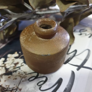 May include: A small, brown, ceramic inkwell with a round opening. The inkwell has a rough, textured surface.