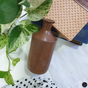 May include: A brown ceramic bottle with a wide mouth and a rounded bottom. The bottle is sitting on a white surface with a green plant and a book in the background.