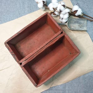 May include: Two rectangular, open-top, red-painted wooden boxes, possibly for storage or display. The boxes are placed on a sheet of brown paper, with a small, weathered metal scoop and cotton sprigs in the background.