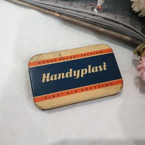 May include: Vintage tin box for Handy Plast First Aid Dressing. The box is blue with white and orange lettering. It says "Handy Pocket Packing" and "Handy Plast" in a stylized font. The box also says "First Aid Dressing".