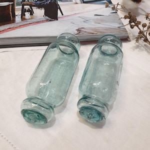May include: Two antique, light blue glass bottles. Each bottle has a cylindrical body, rounded base and top. The bottles have a textured surface and are displayed on a white surface. They are likely decorative or for storing liquids.