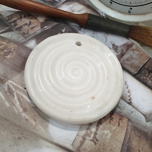 May include: A circular, off-white ceramic pendant with a spiral pattern. It features a small hole for hanging. A brush with a wooden handle is visible in the background.