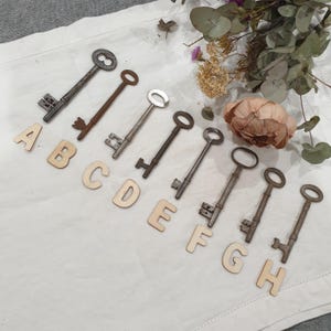 May include: Eight antique keys of various sizes and styles laid out on a white fabric surface with wooden letters spelling out the alphabet from A to H.